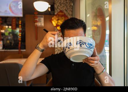 Mann, der im Restaurant in einer Schüssel mit Essstäbchen asiatisches Essen isst. Verschiedene asiatische Gerichte im Restaurant. Asiatisches Speisekonzept Stockfoto