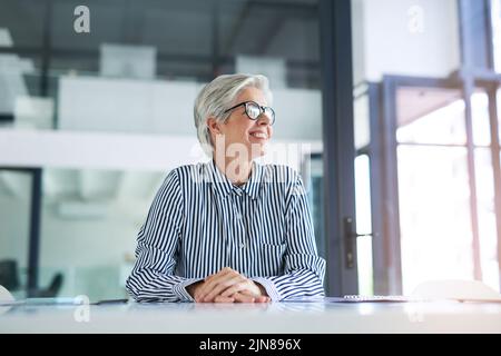 Gerade mit Blick auf die helle Seite des Lebens. Eine attraktive reife Geschäftsfrau, die aus einem Fenster schaut, während sie an ihrem Schreibtisch bei der Arbeit sitzt. Stockfoto