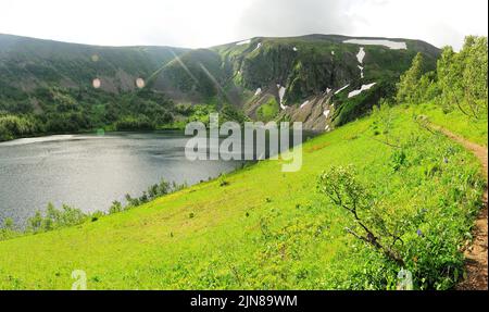 Blick von der Spitze des Hügels im Gras und einem ruhigen See hoch in den Bergen mit den Überresten von Schnee auf den Pisten. Ivanovskie Seen, Chakassien, Stockfoto