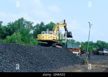 Erdbagger, der auf einem Haufen Kohle stationiert ist, um Kohle in einen Lieferwagen zu laden, wobei zwei andere Lastwagen dabeistehen. Lage: Udipi, Karnataka, Indi Stockfoto