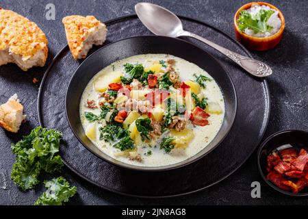 Traditionelle Zuppa Toscana, herzhafte toskanische Suppe mit italienischer Wurst, Grünkohl, gebratenem Speck und Kartoffeln in einer schwarzen Schüssel auf einem grauen Betontisch Stockfoto