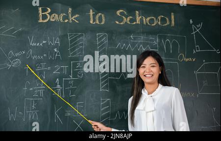 Zurück zum Schulkonzept. Asiatische Lehrerin lächelnd mit Holzstab zeigt auf Tafel in der Schule im Klassenzimmer, glücklich schöne junge Frau Stan Stockfoto