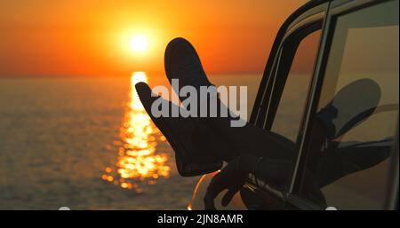 Es ist nur Sie, die Sonne und das Meer. Eine unkenntliche Frau Beine baumeln aus dem Fenster eines Autos am Strand. Stockfoto