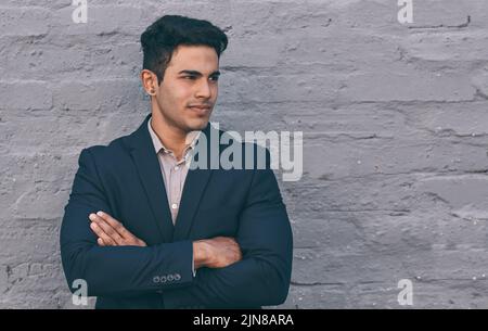 Sein Geist ist auf seinem nächsten Schritt. Ein hübscher junger Geschäftsmann, der mit gefalteten Armen gegen eine Backsteinmauer steht. Stockfoto