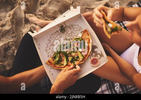 Auf halbem Weg durch unsere Bäuche. Ein junges, nicht erkennbares Paar, das ein kleines Picknick-Date hat und am Strand bei Sonnenuntergang eine Pizza teilt. Stockfoto