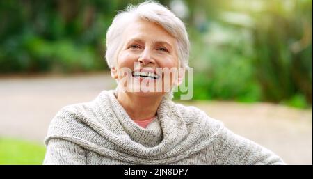 Ich liebe es, Zeit allein zu verbringen. Eine fröhliche ältere Frau sitzt tagsüber auf einer Bank im Park. Stockfoto