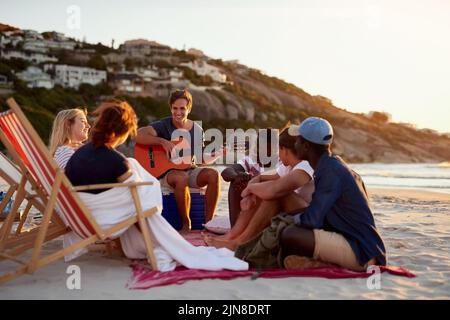 Ein Mann, der Gitarre spielt, während er mit seinen Freunden am Strand sitzt. Stockfoto