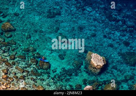 Fotografie von oben von einem nicht wiedererkennbaren erwachsenen Mann, der auf einem blauen SUP Stand-Up Paddleboard zwischen Felsen am türkisfarbenen Ufer steht. Stockfoto