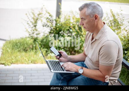 Gealterter grauhaariger Geschäftsmann, der mit einem Smartphone auf einer Bank im grünen Stadtpark sitzt und auf einem digitalen Laptop tippt. Moderner Lebensstil, Fernarbeit, soziale Medien Stockfoto