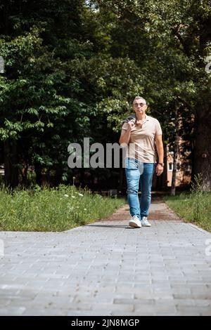 Foto in voller Länge von unten von einem alten brutalen Mann in schwarzer Sonnenbrille, der am sonnigen Tag auf dem grünen Stadtplatz in der Nähe des Haushofs entlang des Pflasterpfades geht. Stadt Stockfoto