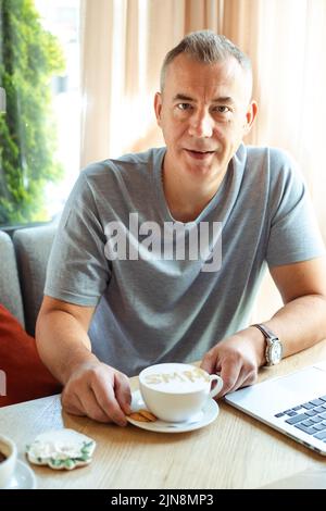 Gealterter, gutaussehender Geschäftsmann, der mit einer Tasse Kaffee im Café sitzt und an einem Laptop arbeitet. Social Media Marketing, SMM, Promotion, Remote Job, gewürzt Stockfoto