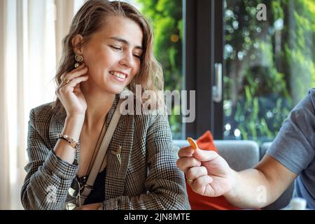 Porträt einer lächelnden jungen Geschäftsfrau in stilvollem Outfit, die am Tisch sitzt und das Gesicht berührt, die Hand des Mannes, der Essen gibt. Urbaner Lebensstil, Ruhe im Café Stockfoto