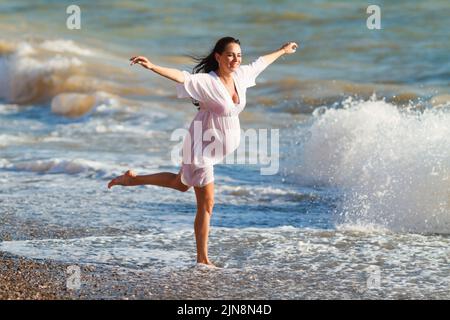 Eine Schwangerin springt am Strand. Eine fröhliche, schwanger Frau rennt an die Küste. Eine Schwangerin in einem romantischen weißen Kleid, die am Strand in der Nähe des tropischen Meeres springt. Indikation von glücklichen Schwangeren. Stockfoto