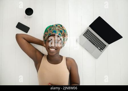 Draufsicht der schönen jungen afrikanischen Frau, die auf weißem Holzboden liegt und einen traditionellen Schal mit Kopfband trägt. .Mit Laptop, Handy und Tasse o Stockfoto