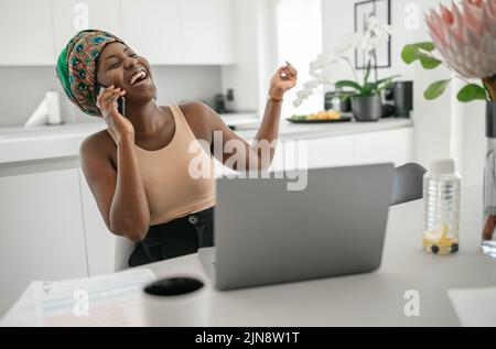 Schöne junge afrikanische Frau trägt Tradition Kopftuch. Sitzen zu Hause und arbeiten auf einem Laptop. Am Telefon lächelnd Stockfoto