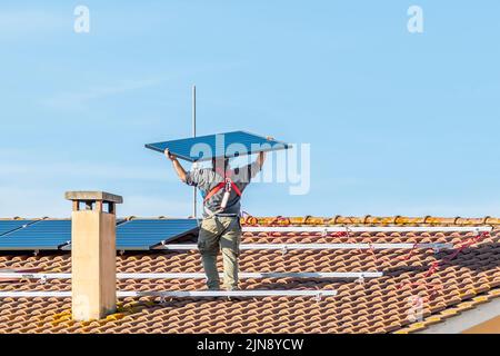 Ein Arbeiter hebt eine Solaranlage auf einem rot gefliesten Dach an, um eine Photovoltaikanlage zu installieren Stockfoto