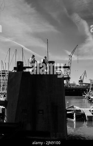 Eine Vertikale von zwei Freunden, die am Hafen von Amsterdam sitzen, wurde in Graustufen gedreht Stockfoto