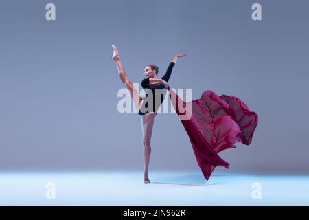 Porträt der jungen flexiblen Ballerina tanzen mit tiefrotem Stoff isoliert über blaugrauen Studio-Hintergrund. Anmutig Stockfoto