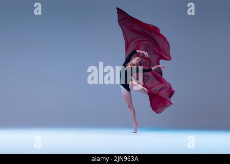 Portrait einer jungen leidenschaftlichen Ballerina, die mit tiefrotem transparentem Stoff auf blaugrauem Studiohintergrund tanzt. Stockfoto