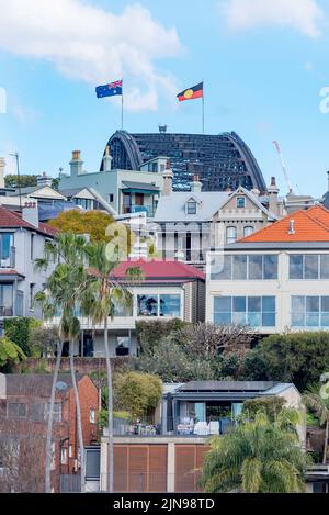 Von Kirribilli aus gesehen, fliegt die Flagge der Aborigines mit starkem Westwind nun neben der australischen Flagge über der Sydney Harbour Bridge Stockfoto