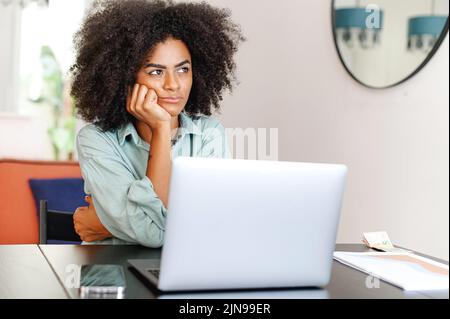 Gelangweilte, müde afroamerikanische Frau, die ihren Laptop für Fernarbeiten benutzt, sitzt im Wohnzimmer, eine Studentin oder Freiberuflerin fühlt sich müde oder interessiert sich nicht für aktuelle Aktivitäten Stockfoto