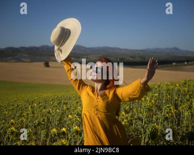 Optimistisches Weibchen in Sonnenbrillen mit Hut in erhobenem Arm, das am Sommertag auf der Plantage mit wachsenden Sonnenblumen auf dem Land steht Stockfoto