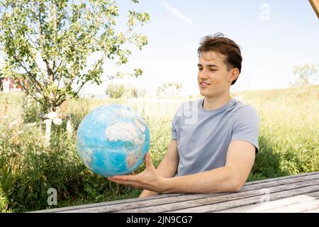 Teenager mit grauem Hemd, der auf einer Parkbank sitzt und den Globus in der Hand hält und von einer Reise, einem Urlaub träumt Stockfoto