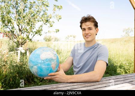 Teenager mit grauem Hemd, der auf einer Parkbank sitzt und den Globus in der Hand hält und von einer Reise, einem Urlaub träumt Stockfoto