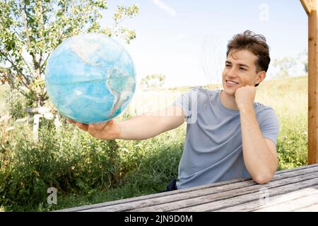 Teenager mit grauem Hemd, der auf einer Parkbank sitzt und den Globus in der Hand hält und von einer Reise, einem Urlaub träumt Stockfoto