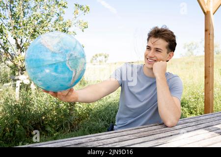 Teenager mit grauem Hemd, der auf einer Parkbank sitzt und den Globus in der Hand hält und von einer Reise, einem Urlaub träumt Stockfoto