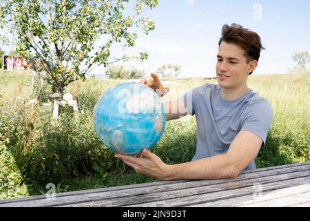 Teenager mit grauem Hemd, der auf einer Parkbank sitzt und den Globus in der Hand hält und von einer Reise, einem Urlaub träumt Stockfoto