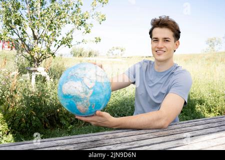 Teenager mit grauem Hemd, der auf einer Parkbank sitzt und den Globus in der Hand hält und von einer Reise, einem Urlaub träumt Stockfoto