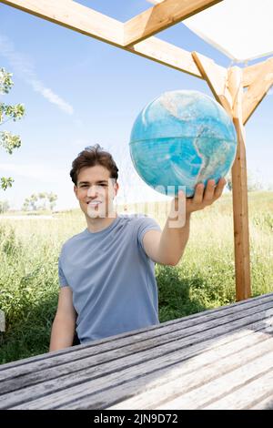 Teenager mit grauem Hemd, der auf einer Parkbank sitzt und den Globus in der Hand hält und von einer Reise, einem Urlaub träumt Stockfoto