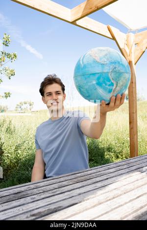 Teenager mit grauem Hemd, der auf einer Parkbank sitzt und den Globus in der Hand hält und von einer Reise, einem Urlaub träumt Stockfoto