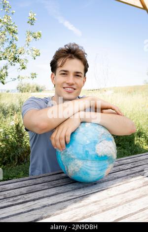 Teenager mit grauem Hemd, der auf einer Parkbank sitzt und den Globus in der Hand hält und von einer Reise, einem Urlaub träumt Stockfoto