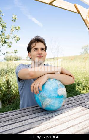 Teenager mit grauem Hemd, der auf einer Parkbank sitzt und den Globus in der Hand hält und von einer Reise, einem Urlaub träumt Stockfoto