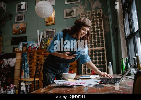 Junge Frau benutzt Handy und Laptop während sie in ihrem Studio arbeitet Stockfoto