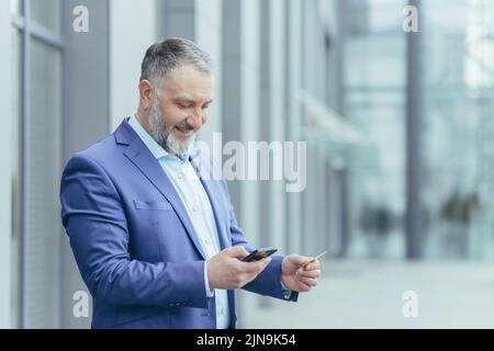 Der ältere grauhaarige Geschäftsmann vor dem Bürogebäude, der Mann im Geschäftsanzug nutzt das Mobiltelefon-Smartphone, um im Online-Shop und im Bestellservice zu kaufen, hält die Bankkarte in den Händen Stockfoto