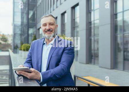 Porträt eines erfolgreichen älteren grauhaarigen Geschäftsmanns-Chefs, eines Mannes außerhalb des Büros mit einem Tablet-Computer, der lächelt und die Kamera anschaut, eines Investors im Geschäftsanzug, eines glücklichen Bankiers Stockfoto