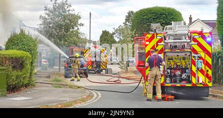 Feuerwehrmänner von Essex Fire and Rescue Service sprühen Wasserstrahl auf Haus zwei Feuerwehrfahrzeuge und Feuerwehrleute in Wohnstraßenlandschaft England Großbritannien Stockfoto