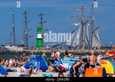 Rostock, Deutschland. 10. August 2022. Vor dem gut besuchten Strand segeln historische Segelschiffe am Pier entlang in Richtung Stadthafen. Das sonnige und warme Wetter wird von Urlaubern und Tagesbesuchern für Strandausflüge an der Ostseeküste genutzt. Quelle: Jens Büttner/dpa/Alamy Live News Stockfoto