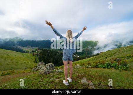 Eine junge, schlanke, blonde Frau in Shorts und einem Hemd steht mit erhobenen Händen auf einem Berg über den Wolken. Bakhmaro, Georgien Stockfoto