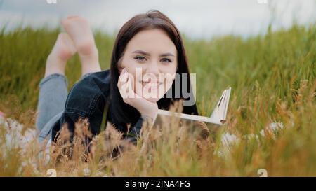 Junge heitere kaukasische Brunet Frau Mädchen auf Gras im Freien liegend Attraktive lächelnde Teenager-Student liest Buch genießen romantische Literatur Geschichte entspannen Stockfoto