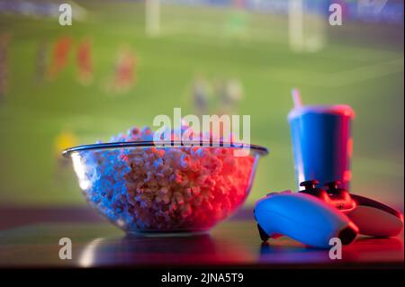 Fußballspiel auf dem Bildschirm eines großen Plasmafernsehens. Popcorn in einer Glasschüssel, ein kohlensäurehaltiges Getränk in einem Plastikglas und ein Gamepad. Fastfood-Videospiele Stockfoto