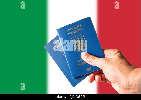 Vor dem Hintergrund der italienischen Nationalflagge zwei ukrainische biometrische Pässe in der Hand einer Frau. Auswanderung der Ukrainer nach Italien, Arbeit, tou Stockfoto