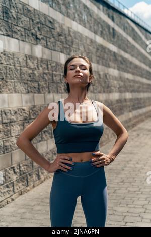 Die sportliche Frau trägt Trainingsausrüstung, die Hände liegen auf der Hüfte und die Augen geschlossen Stockfoto