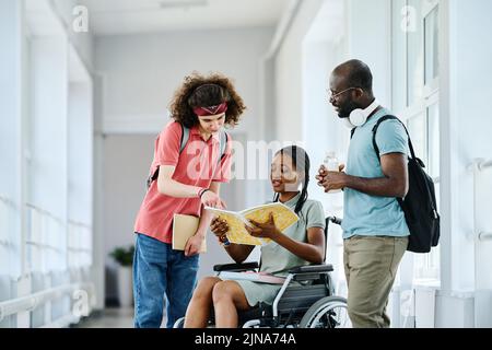 Gruppe multiethnischer Studenten, die ein Lehrbuch lesen und es gemeinsam diskutieren, während sie in der Pause am Korridor stehen Stockfoto