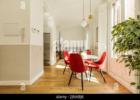 Wohnzimmer in der Wohnung mit Esstisch mit roten Samtpolsterstühlen, Balkon mit weißen Holztüren und Kastanienboden Stockfoto