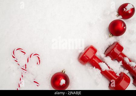 Kurzhanteln, Weihnachtskugeln-Ornamente, Zuckerrohr auf weißem Schnee. Fitness-Studio-Komposition, Training bei kaltem Winterwetter, flach liegend mit Kopierfläche. Stockfoto