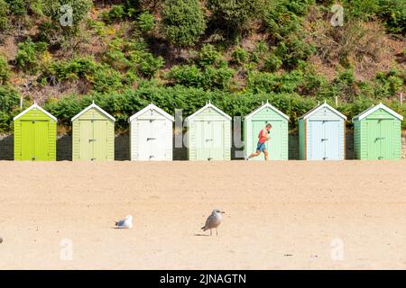 Eine Reihe von mehrfarbigen Strandhütten aus Holz und einem männlichen Läufer, der am Meer in Bournemouth Dorset, Großbritannien, läuft Stockfoto